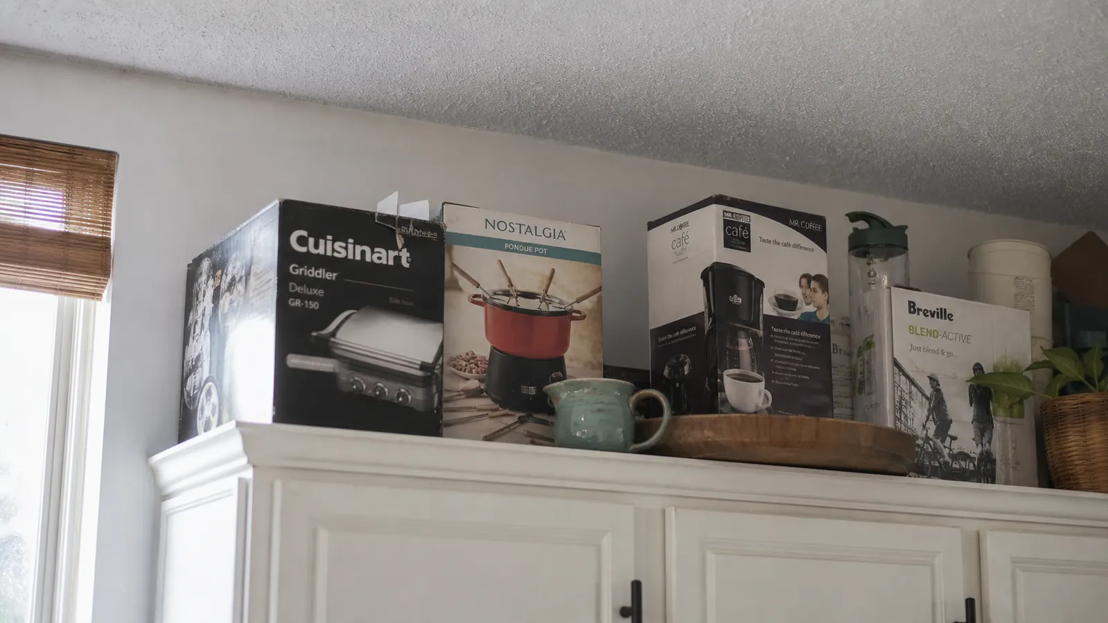 A high cabinet shelf with the fondue pot box tucked between other kitchen appliances