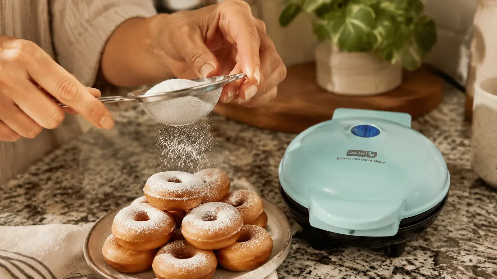 Hands sprinkling powdered sugar over a plate of fresh mini donuts next to the Dash mini donut maker on a kitchen counter