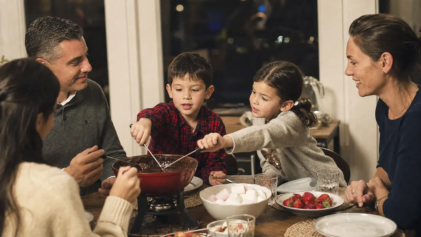 Grandchildren laughing while dipping forks into a chocolate fondue pot with strawberries and marshmallows nearby